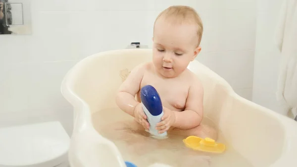 Küçük bebek banyoda yıkanıyor ve elinde şampuanla ya da duş jeliyle şişe tutuyor. — Stok fotoğraf