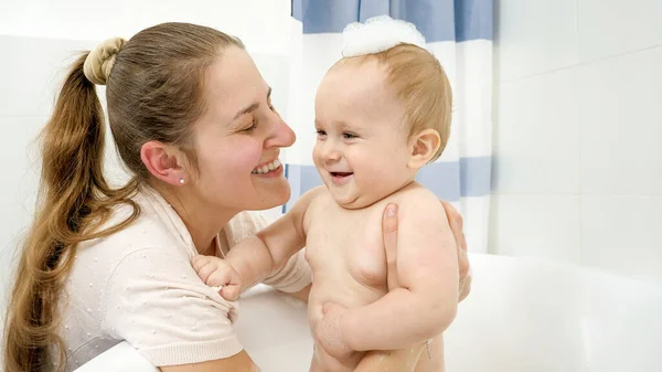 Retrato de niño sonriente codiciado en espuma de jabón y madre lavándolo en el baño — Foto de Stock