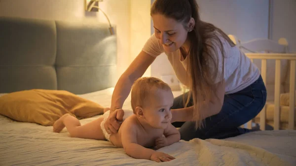 Caring mother putting on diapers on her little baby son crawling on bed at night. Concept of children hygiene, healthcare and family care at home — Stock Photo, Image