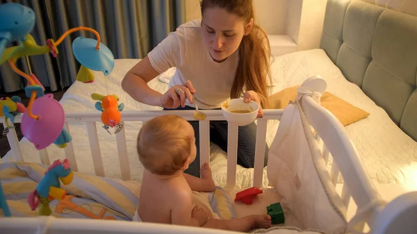 Молодая заботливая мать кормит своего маленького сына супом в детской кроватке ночью. Концепция здорового питания, воспитания и ухода за детьми — стоковое фото