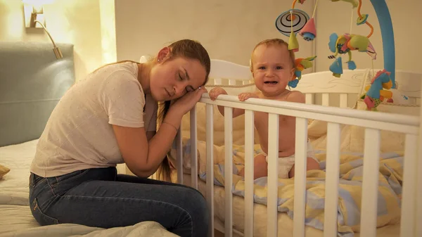 Pequeño niño gritando y llorando junto a su madre cansada durmiendo junto a su cuna. Concepto de paternidad, fatiga de los padres y desarrollo de los niños. —  Fotos de Stock