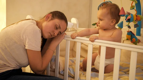 Lindo bebé niño sentado en la cuna y mirando a dormir madre cansada —  Fotos de Stock