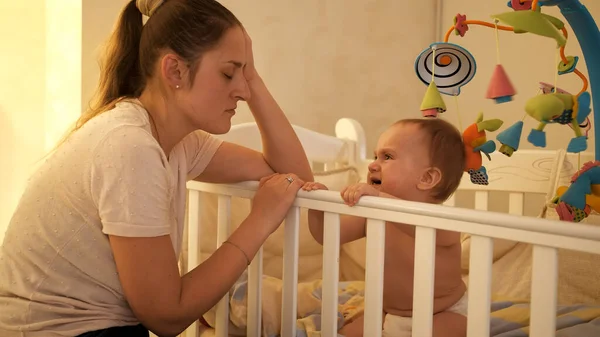 Cansada mãe sonolenta balançando berço de seu bebê chorando e se recusando a dormir à noite. Conceito de parentalidade, fadiga dos pais e desenvolvimento das crianças. — Fotografia de Stock