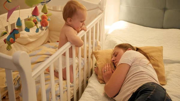 Молодая уставшая мать заснула рядом со своим ребенком, плачущим в постели. Концепция воспитания, усталости родителей и развития детей. — стоковое фото