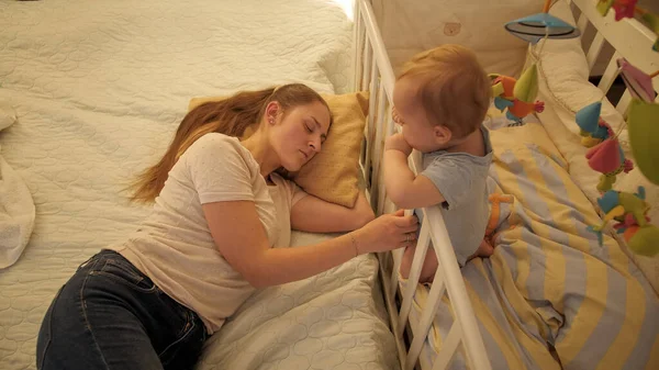 Unavená vyčerpaná matka houpe dětskou postýlkou a spí. Koncepce rodičovství, únavy rodičů a vývoje dětí. — Stock fotografie