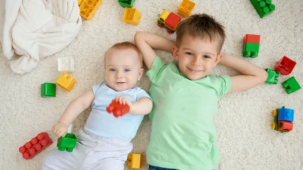Portret uśmiechniętego chłopca ze starszym bratem bawiącym się w klocki i klocki podczas leżenia na dywanie. Koncepcja rozwoju, edukacji i kreatywności dzieci w domu — Zdjęcie stockowe