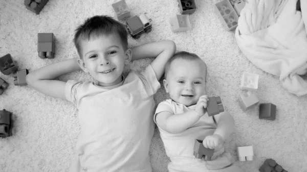 Imagem em preto e branco de menino sorridente alegre com irmão bebê cercado de brinquedos deitados no tapete na sala de jogos. Conceito de desenvolvimento infantil, educação e criatividade em casa — Fotografia de Stock