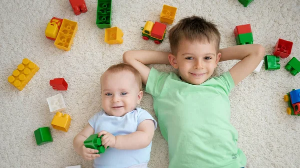 Uśmiechnięty chłopczyk ze starszym bratem leżącym na dywanie obok sterty kolorowych zabawek i patrzącym w górę przed kamerą. Koncepcja rozwoju, edukacji i kreatywności dzieci w domu — Zdjęcie stockowe