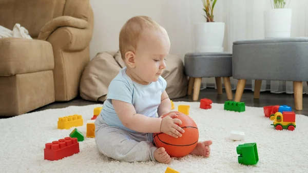 Счастливый улыбчивый мальчик, сидящий на ковре и играющий дома с баскетбольным мячом. Концепция развития детей, спорта, образования и творчества на дому — стоковое фото