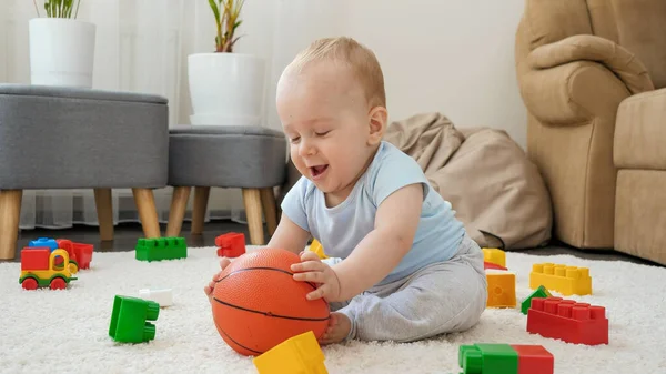 Веселый улыбающийся мальчик веселится и играет с баскетболом на ковре дома — стоковое фото
