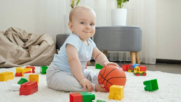Cute uśmiechnięty chłopiec siedzi na dywanie i trzyma piłkę do kosza — Zdjęcie stockowe
