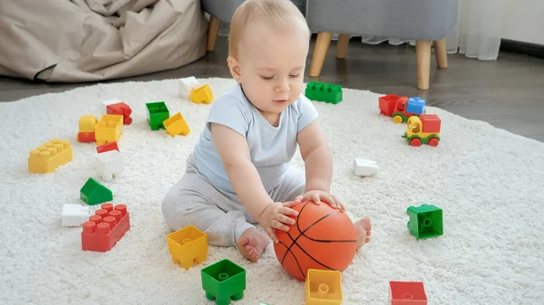 Piccolo bambino che rotola e sbatte pallone da basket sul tappeto a casa. Concetto di sviluppo dei bambini, sport, educazione e creatività a casa — Foto Stock