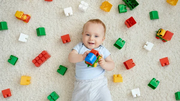 Glad leende pojke med massor av leksaker som ligger på mattan och tittar upp i kameran. Begreppet barns utveckling, utbildning och kreativitet i hemmet — Stockfoto
