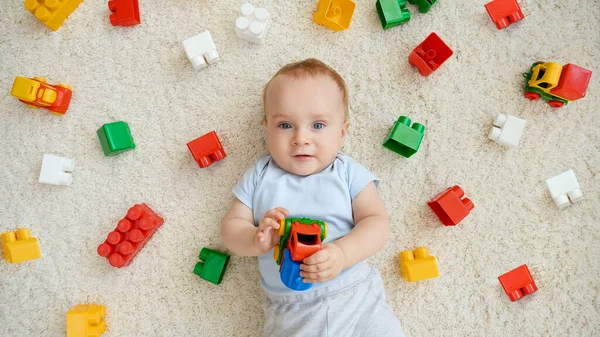 Mosolygó kisfiú portréja, körülvéve játékokkal, amik a szőnyegen fekszenek a játszószobában. A gyermekek fejlődésének, oktatásának és kreativitásának fogalma otthon — Stock Fotó