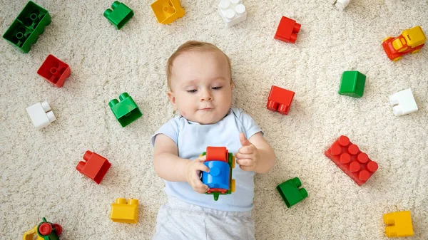 Милый мальчик, играющий в игрушечную машинку на ковре рядом с кучей разноцветных игрушек, блоков и кирпичей. Концепция развития детей, образования и творчества на дому — стоковое фото