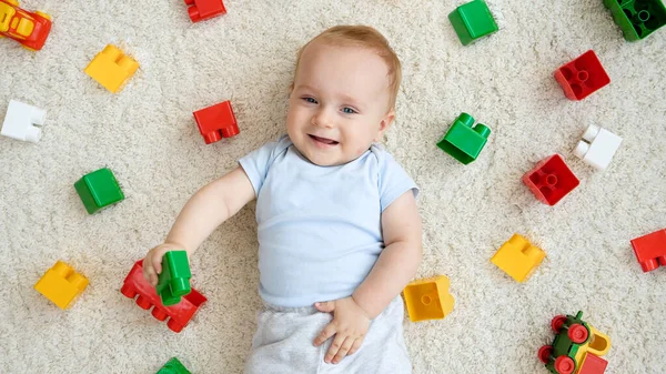 Glad leende liten pojke ligger på mattan täckt med färgglada leksaker, block och tegelstenar. Begreppet barns utveckling, utbildning och kreativitet i hemmet — Stockfoto