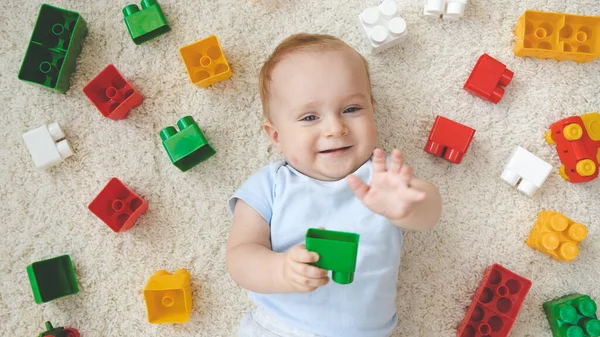 Buon bambino sorridente sdraiato su un tappeto ricoperto di giocattoli colorati, blocchi e mattoni. Concetto di sviluppo dei bambini, educazione e creatività a casa — Foto Stock