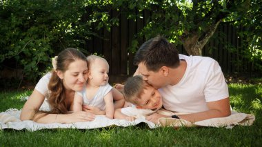 Mutlu genç ebeveynler çocuklarını kucaklıyor ve öpüyor bahçedeki çimlerin üzerinde yatıyorlar.