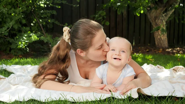 Szczęśliwy uśmiech dziecka leżącego na trawie, podczas gdy matka całuje go w głowie — Zdjęcie stockowe