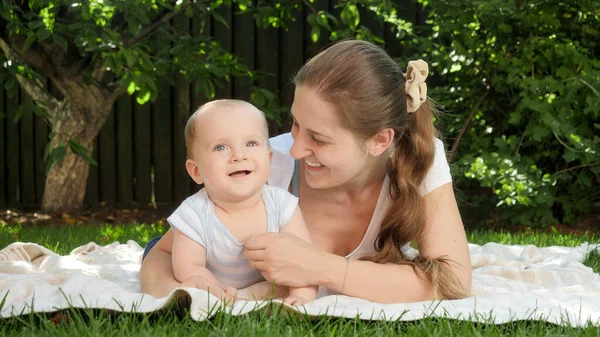 Schattige baby jongen en moeder glimlachend in camera terwijl ze op groen gras liggen in tuin of park. Ouderschap, familie, ontwikkeling van kinderen en plezier in de natuur. — Stockfoto