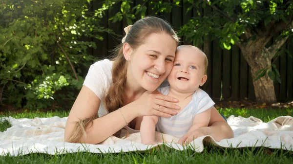 Mooie glimlachende vrouw picknicken met haar kleine baby zoon in het park en knuffelen hem op gras — Stockfoto