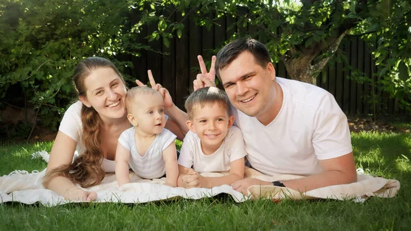 Felice genitori sorridenti sdraiati sull'erba e fare orecchie o v-segno con le dita sulla foto ai loro figli — Foto Stock