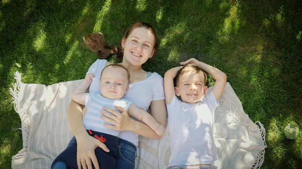 Vista superior de la madre sonriente con dos hijos acostados sobre una manta en la hierba y mirando al cielo. Crianza, familia, desarrollo infantil y diversión al aire libre en la naturaleza. — Foto de Stock