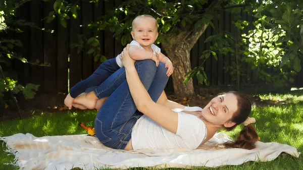 Feliz sorrindo e rindo mãe com o bebê filho brincando no jardim enquanto deitado na grama. Paternidade, família, desenvolvimento infantil e diversão ao ar livre na natureza. — Fotografia de Stock