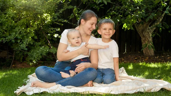 Старший мальчик обнимает младших брата и мать, сидящих на траве в саду. Общение, семья, развитие детей и развлечения на природе. — стоковое фото