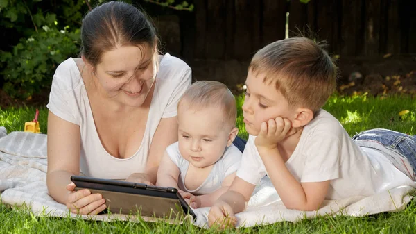 Gelukkige lachende jongen met broertje en moeder liggend op gras in het park en surfen op internet op tablet computer. Ouderschap, gezin, ontwikkeling van kinderen en technologie — Stockfoto