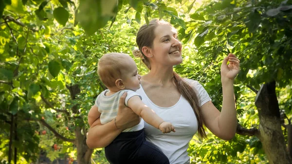 Feliz mãe sorridente com bebê menino andando e realxing no jardim da árvore da maçã no dia ensolarado — Fotografia de Stock