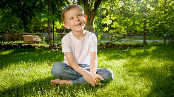 Netter lächelnder Junge, der auf Gras im Hinterhofgarten sitzt und in die Kamera lächelt. Kindererziehung, Familie, Entwicklung der Kinder und Spaß in der Natur. — Stockfoto
