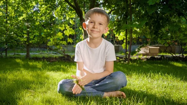 Dolly-Aufnahme eines 6-jährigen Jungen, der auf Gras sitzt und in die Kamera schaut. Kindererziehung, Familie, Entwicklung der Kinder und Spaß in der Natur. — Stockfoto