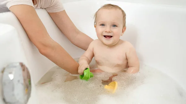 Anne oğlunu destekliyor ve kucağında tutuyor banyoda oturuyor ve oyuncaklarla oynuyor. Evde çocuk hijyeni, sağlık ve geliştirme kavramı — Stok fotoğraf