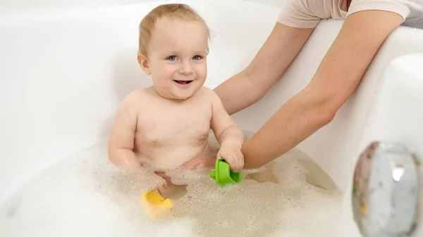 Glücklich lächelnder kleiner Junge, der mit Spielzeug spielt und in der Badewanne mit Wasser spritzt. Konzept der Kinderhygiene, -gesundheit und -entwicklung zu Hause — Stockfoto