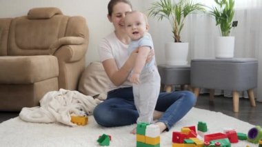 Mutlu gülümseyen anne küçük oğlunu destekliyor ve oturma odasında halıda yürümeyi öğretiyor. Çocuk gelişimi, eğitim ve yaratıcılık kavramı
