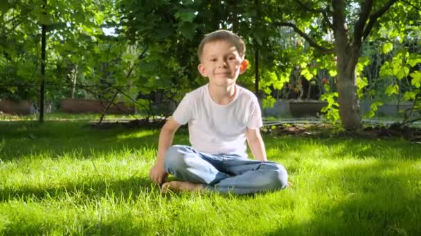6 yaşındaki bir çocuğun çimlere oturup kameraya baktığı Dolly çekimi. Ebeveynlik, aile, çocuk gelişimi ve doğada eğlence.. — Stok video
