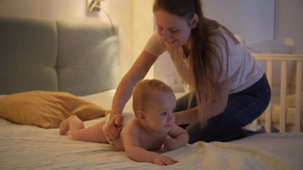 A gondoskodó anya pelenkát vesz fel a kisfiára, aki éjszaka mászik az ágyon. A gyermekhigiénia, az egészségügyi ellátás és a családi gondozás fogalma otthon — Stock videók