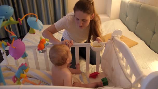 Jonge zorgzame moeder voedt haar zoontje 's nachts met soep in babybedje. Concept van gezonde voeding, ouderschap en babyverzorging — Stockvideo