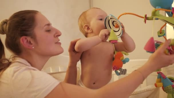 Bonito menino e mãe brincando com brinquedos de fiação coloridos no berço do bebê. Conceito de parentalidade, desenvolvimento infantil. — Vídeo de Stock