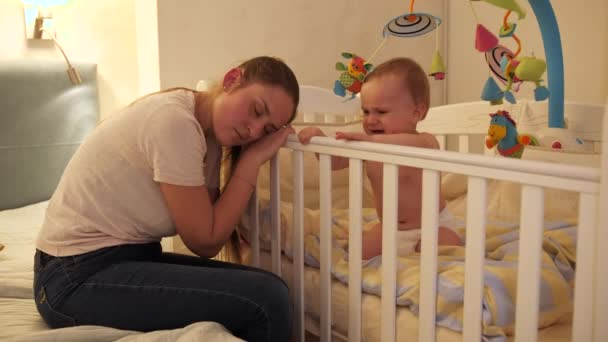 Pequeño niño gritando y llorando junto a su madre cansada durmiendo junto a su cuna. Concepto de paternidad, fatiga de los padres y desarrollo de los niños. — Vídeo de stock