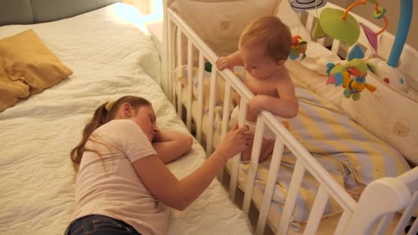 Mały bezsenny chłopczyk kołyszący się w łóżeczku obok zmęczonej matki śpiącej w łóżku. Pojęcie rodzicielstwa, zmęczenia rodziców i rozwoju dzieci. — Wideo stockowe