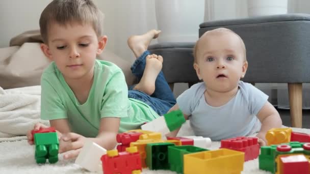 Niño pequeño con hermano mayor jugando bloques de juguete y ladrillos en la alfombra en la sala de estar. Concepto de desarrollo infantil, educación y creatividad en el hogar — Vídeos de Stock
