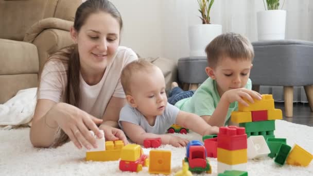 Feliz familia sonriente con el bebé jugando en la alfombra con bloques de juguete, ladrillos y coches. Concepto de familia teniendo tiempo juntos y desarrollo de los niños — Vídeo de stock