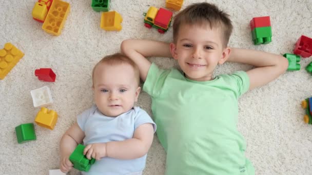 Alegre niño sonriente con hermano pequeño rodeado de juguetes acostados en la alfombra en la sala de juegos. Concepto de desarrollo infantil, educación y creatividad en el hogar — Vídeos de Stock