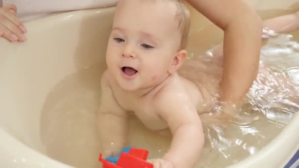 Мама поддерживает своего маленького сына бледнеющего и купающегося в пластиковой ванне дома. Концепция детской гигиены, здравоохранения и воспитания. — стоковое видео