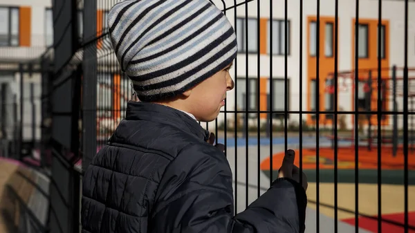 Mały zdenerwowany chłopiec spoglądający przez metalowy płot na nowoczesną szkołę i plac zabaw. Pojęcie ubóstwa, imigracji, znęcania się i stresu wśród dzieci — Zdjęcie stockowe