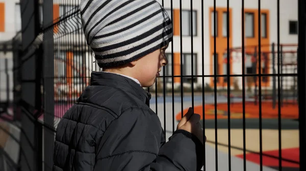 Zdenerwowany samotny chłopiec patrzący przez metalowy płot na dziecięcy placyk i nowoczesną szkołę — Zdjęcie stockowe