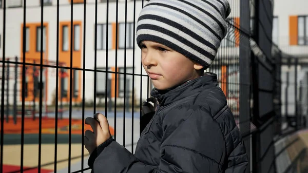 Maltratado y estresado niño pequeño sosteniendo valla de metal y mirando a los niños palyground — Foto de Stock