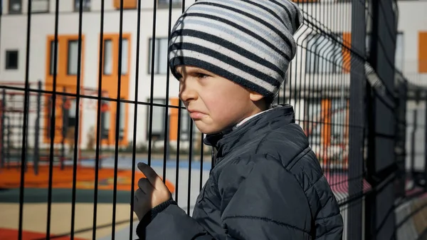 Molesto niño solitario lenaing y la celebración de cerca de metal red en el patio de recreo de la escuela — Foto de Stock